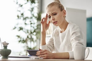 Image of a female sitting at a table looking worried and holding a calculator in one hand and the other hand is holding her head.