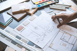 An Interior Designer's desk on top of which are drawings of a living space, samples for colours and furniture finishes as well as different coloured paints and pens. 