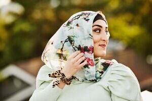 A woman is wearing a light blue patterned headscarf and a chic blue blouse with a trees in the background.