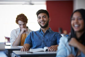 Male student smiling in classroom with other classmates with book and mobile phone on the table